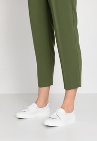Des jambes portant un pantalon cropped olive et des baskets blanches à enfiler avec un détail de nœud torsadé, debout sur un sol clair.