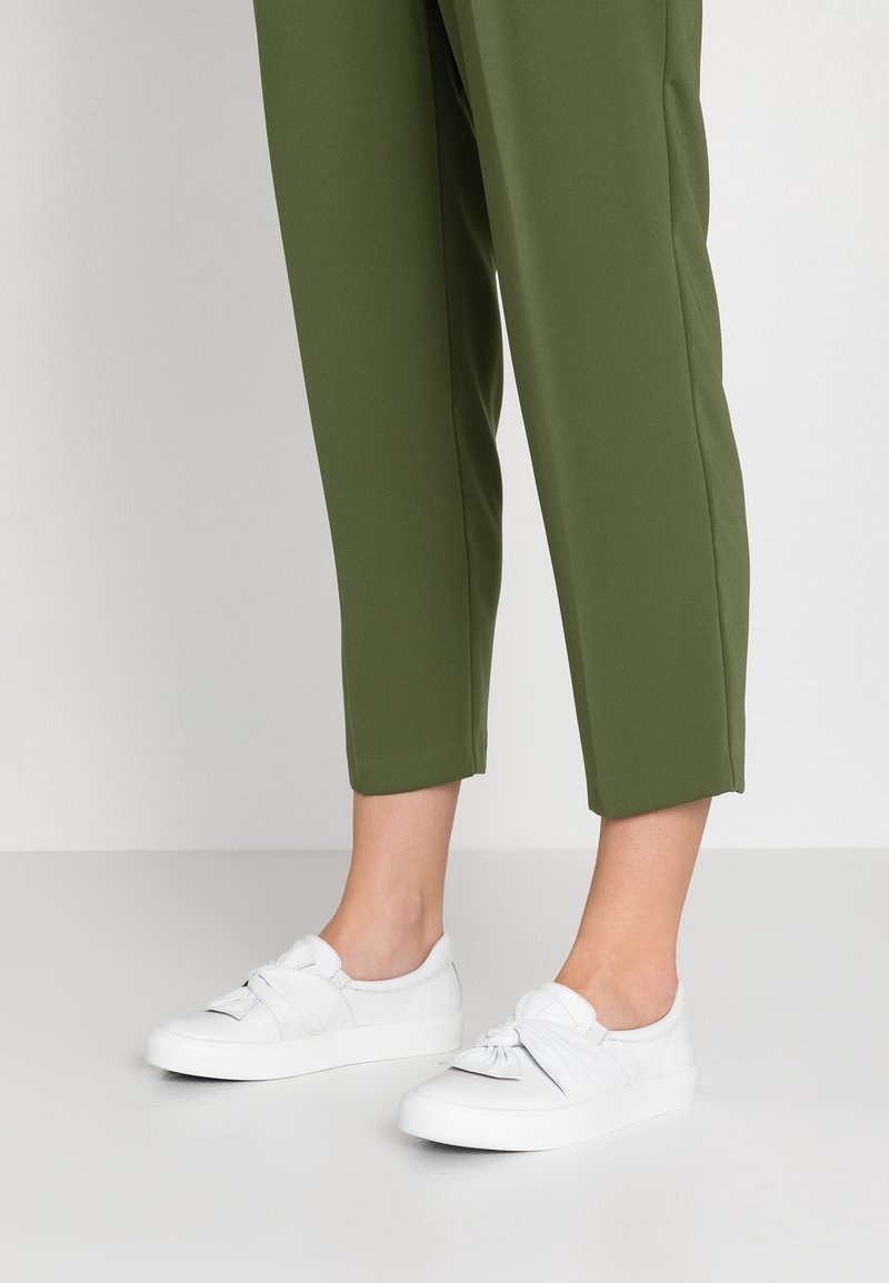 Des jambes portant un pantalon cropped olive et des baskets blanches à enfiler avec un détail de nœud torsadé, debout sur un sol clair.