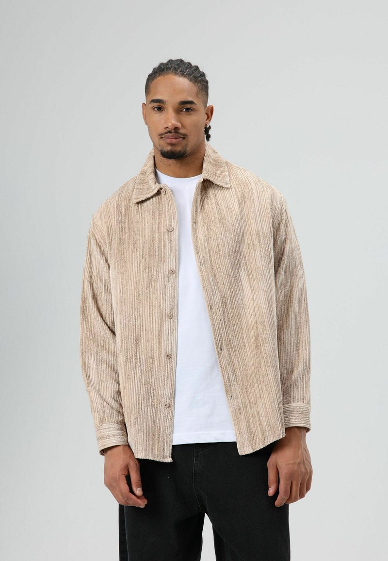 Licht beige corduroy overhemd met een knoopsluiting aan de voorkant en een spitse kraag, gedragen over een wit T-shirt, afgebeeld tegen een neutrale achtergrond.