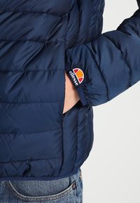 Námorní modrá péřová bunda s prošívanou texturou, boční kapsou a oranžovým, bílým a námořnickým logem na manžetě rukávu. Odolná tkanina.