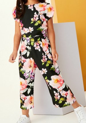 Combinaison noire à fleurs avec des manches courtes bouffantes, des fleurs roses et blanches vives, des jambes cropped et un tissu doux. Associée à des baskets blanches.