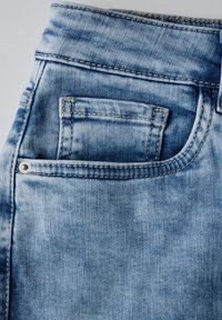 Světle modré džíny z denimu s zadní kapsou, oranžovým prošíváním a kovovým nýtem jako akcentem. Tkanina vykazuje opotřebovaný vzhled.