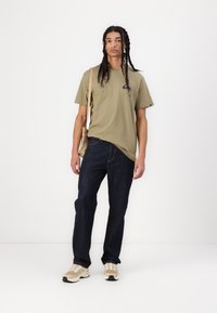 Olivengrønn T-skjorte med logo, mørkeblå denimjeans, beige sneakers med svarte detaljer, og en lys tan crossover-veske.