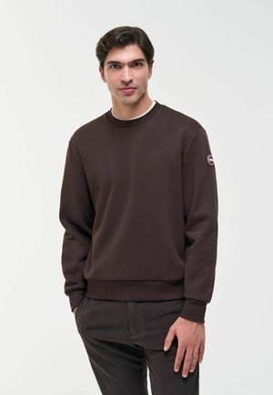 Giovane uomo con capelli scuri che indossa una felpa marrone e pantaloni scuri, in piedi davanti a uno sfondo chiaro e semplice.