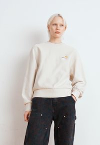 Beige bomulds sweatshirts med ribbede manchetter og kant, der har et lille broderet logo i gul og sort på brystet.