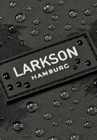 Material textil negru cu picături de apă, având un eticheta rectangulară neagră cu text alb „LARKSON HAMBURG” și studuri decorative.