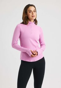 Top atletico a maniche lunghe di un rosa chiaro, con collo a zip a un quarto e aperture per i pollici, abbinato a leggings neri. Texture liscia e design aderente.