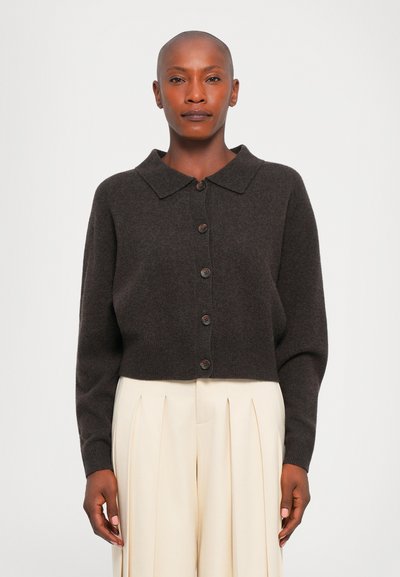 Personne portant un cardigan boutonné chocolat avec un col et un pantalon crème plissé taille haute, se tenant devant un fond uni.