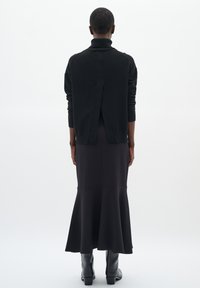 Maglione nero con colletto alto e una fessura sul retro, abbinato a una gonna a trapezio nera aderente. Indossato con stivaletti neri alla caviglia.