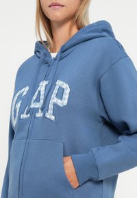 Blå hettegenser med en teksturert "GAP"-logo i lyseblått, snorhette og frontlommer. Myk stoffkvalitet og avslappet passform.
