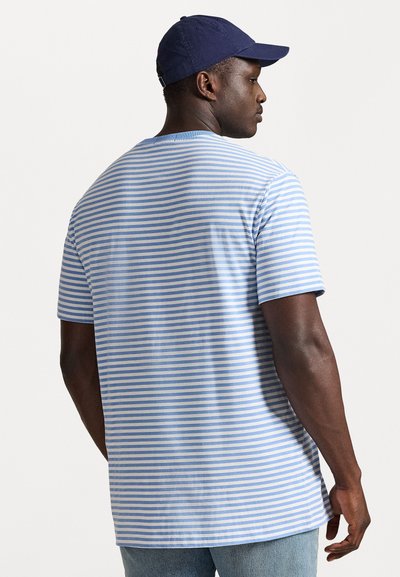 Gaiš zilas un baltas svītrainas īsās piedurknes T-krekls, izgatavots no kokvilnas, ar brīvu piegriezumu un apaļu kakla izgriezumu, uzvilkts ar tumši zilu cepuri.