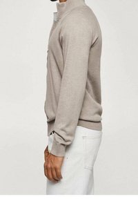 Maglione beige in maglia con colletto alto e design a mezza zip. Presenta una silhouette aderente e polsini a coste, abbinato a pantaloni bianchi.