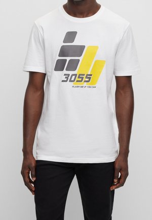 Mann trägt ein weißes T-Shirt mit grauen und gelben geometrischen Formen, der Zahl "3055" und dem Text "FLASH ME IF YOU CAN" auf der Vorderseite.