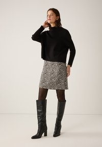 Schwarzer, kurzer Pullover, gemusterter Minirock mit beige- und schwarzem Design, durchsichtige, gemusterte Strumpfhosen und kniehohe schwarze Lederstiefel.