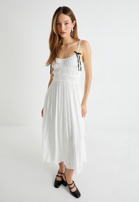 Witte jurk met een gesmokte bodice, dunne bandjes met zwarte strikjes, en een vloeiende asymmetrische zoom. Gecombineerd met zwarte blok hakken.