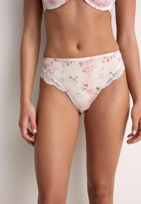 Sieviete valkā gaiši rozā ziedu augsti izgrieztas biksītes ar mežģīņu apdari, redzama no vidukļa līdz vidējai augšstilbu daļai pret vienkāršu fonu.