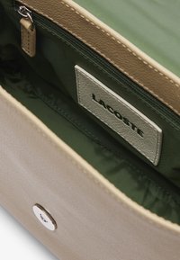 Intérieur d'un sac à main en cuir beige avec une doublure textile verte, comprenant une poche zippée et une étiquette avec "LACOSTE" en noir. Détail du fermoir rond visible.