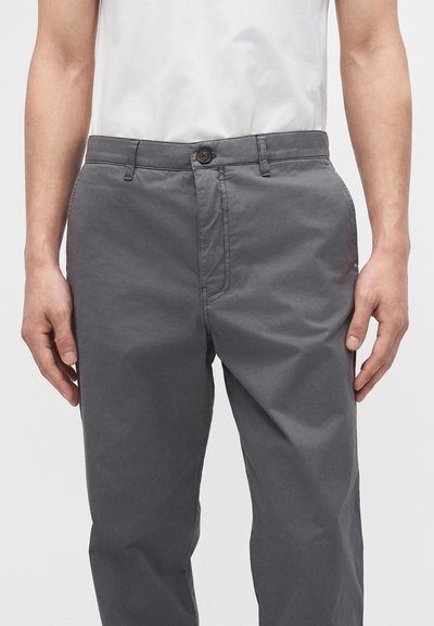 Homme portant un pantalon décontracté gris avec bouton et fermeture éclair à l'avant, associé à une chemise blanche, debout, les mains détendues le long du corps.