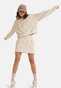 Beige cropped hoodie with front pocket, paired with a beige mini skirt featuring side pockets. White sneakers and pink socks complete the look.