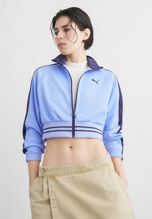 Giovane con capelli corti e scuri che indossa una giacca corta Puma azzurro chiaro e una gonna beige, con le mani in tasca, che guarda di lato.