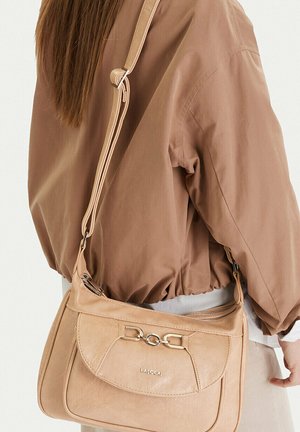 Osoba w brązowej kurtce niosąca beżową torbę na ramię marki Lasocki z metalową złotą klamrą i regulowanym paskiem.