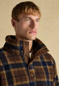 Brown and blue plaid fleece pullover with a high collar and snap button closure. Soft texture with contrasting brown fabric accents.