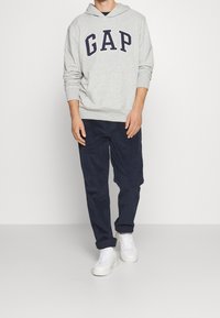 Felpa grigia con logo "GAP" in blu navy, sneakers bianche e pantaloni blu scuro. Il tessuto sembra morbido e informale, adatto per l'uso quotidiano.