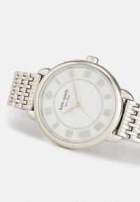 Silverfärgad metallarmbandsur med en vit urtavla, romerska siffror, guldfärgade visare och ett strukturerat nätarmband. Har en liten spadlogo.