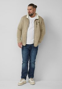 Beige Fleecejacke mit Reißverschluss vorne, weißem Kragen und Brusttasche, kombiniert mit blauen Jeans und weißen Sneakern. Darunter ein schlichtes weißes T-Shirt.