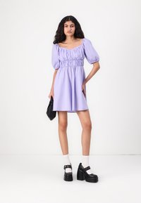 Robe courte lilas avec des manches bouffantes, un corsage froncé et une jupe évasée. Accompagnée de chaussures à plateforme noires et d'un petit sac à main noir.