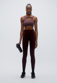 Top sportivo corto color malva con dettaglio a taglio, abbinato a leggings bordeaux a vita alta. Tiene una borraccia nera, indossa sneakers nere.