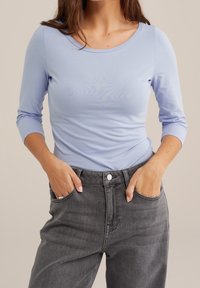 Ljusblå långärmad topp med rundad halsringning, figursydd kropp och slät textur, kombinerad med högmidjade mörkgrå jeans.