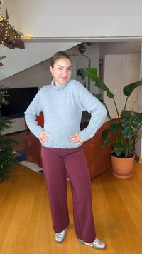 Pull gris en maille avec un col montant, associé à un pantalon large bordeaux. Des baskets argentées complètent la tenue ; cadre intérieur avec des plantes.
