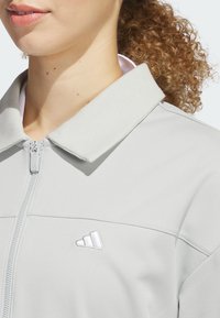 Ljusgrå zip-up jacka med ribbad krage och broderad logotyp. Har en slät yta och en figurnära design som visar en modern stil.