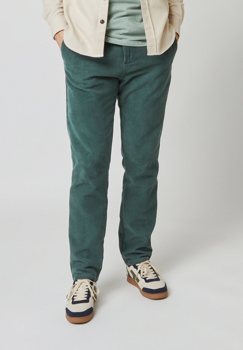 Groene corduroybroek met een slim fit, voorzien van verticale ribbels en gecombineerd met crèmekleurige en marineblauwe sneakers. De stof lijkt gestructureerd.