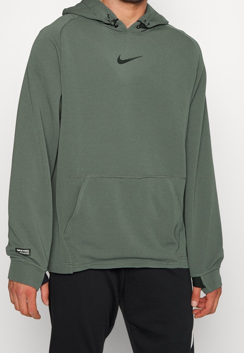Homme portant un sweat à capuche Nike vert olive avec poche avant kangourou et étiquette Nike Pro noire sur la manche, assorti à un pantalon noir.