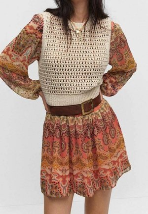 Vrouw die een lange jurk met een paisley-patroon draagt, met een gebreid sleeveless crème vest en een bruine riem met een vierkante gesp.