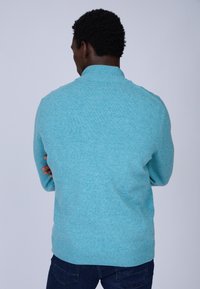Pull en maille turquoise avec col montant, tissu texturé, poignets et ourlet côtelés, vu de dos. Associé à un jean en denim foncé.
