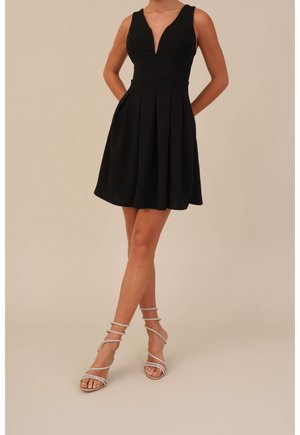 Kobieta w czarnej sukience bez rękawów z głębokim dekoltem w kształcie litery V i rozkloszowaną spódnicą, w połączeniu ze srebrnymi sandałami na wysokim obcasie z paskami.