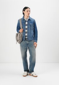 En ung mann står, iført en denimjakke, stripete skjorte og jeans, og holder en grå skulderveske med en enkel hvit bakgrunn.