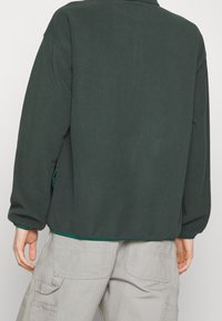 Mørkegrøn sweatshirt med en afslappet pasform, der har lange ærmer, ribstrikkede manchetter og en kontrasterende grøn kant. Blød tekstur, ingen synlige mønstre.