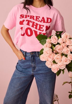 Nainen pukeutuneena vaaleanpunaiseen t-paitaan, jossa on punainen "Spread the Love"-teksti, pitää suurta kimppua vaaleanpunaisia ruusuja vaaleanpunaisen taustan edessä.