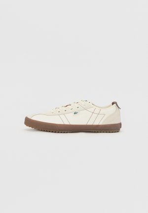 Zapatillas de hombre de lona crema con suela de goma marrón, que presentan detalles de costura sutiles y un pequeño logo verde en el lateral.