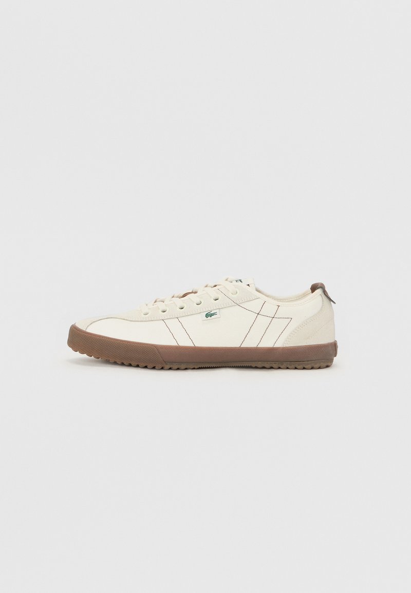 Zapatillas de hombre de lona crema con suela de goma marrón, que presentan detalles de costura sutiles y un pequeño logo verde en el lateral.