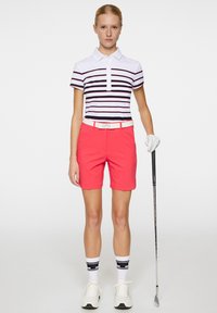 Golfoutfit med en vit och marinblå randig polo, korallfärgade shorts, vit bälte och vita sportskor med randiga strumpor, hållande en klubba.