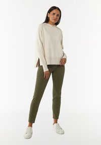 Jasno beżowy sweter dzianinowy z wysokim kołnierzem i bocznymi rozcięciami, w połączeniu z oliwkowo zielonymi spodniami slim-fit i białymi trampkami.