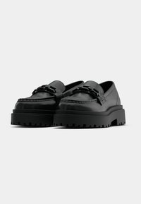 Mocassins de couro preto com sola baixa tipo plataforma, apresentando um detalhe de fivela metálica e costura ao longo da borda superior.