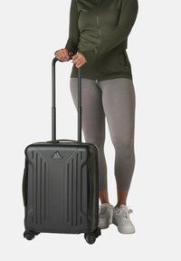 Hartschaliger schwarzer Kabinenkoffer mit vertikalen Rillen, ausziehbarem Griff, vier Rollen und einem gut sichtbaren Logo auf der Vorderseite.