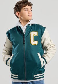 Teal and cream varsity jacket with a letter "C" patch, zippered front, ribbed cuffs, and two front pockets. Layered over a gray hoodie.