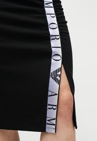 Μαύρη φούστα με πλευρική σχισμή που έχει μια λευκή κάθετη ρίγα με το κείμενο "Emporio Armani" και το λογότυπο κατά μήκος ενός γυμνού ποδιού.